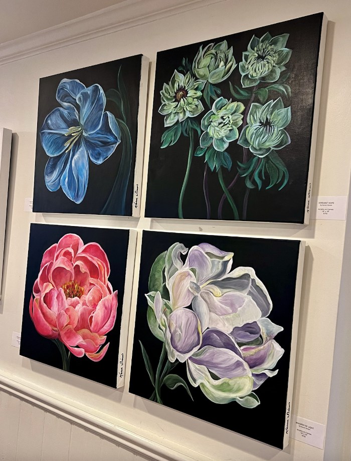 Four of Karen Simon’s floral paintings on black backgrounds — a blue tulip, green hellebores, a pink peony, and a white-and-lavender bloom — grouped on a gallery wall.