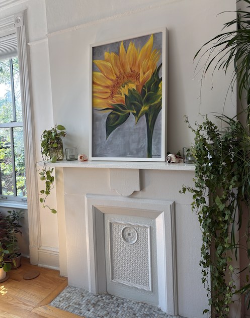 Yellow sunflower painting by Karen Simon in a white frame, hung above a white fireplace mantel with trailing houseplants.